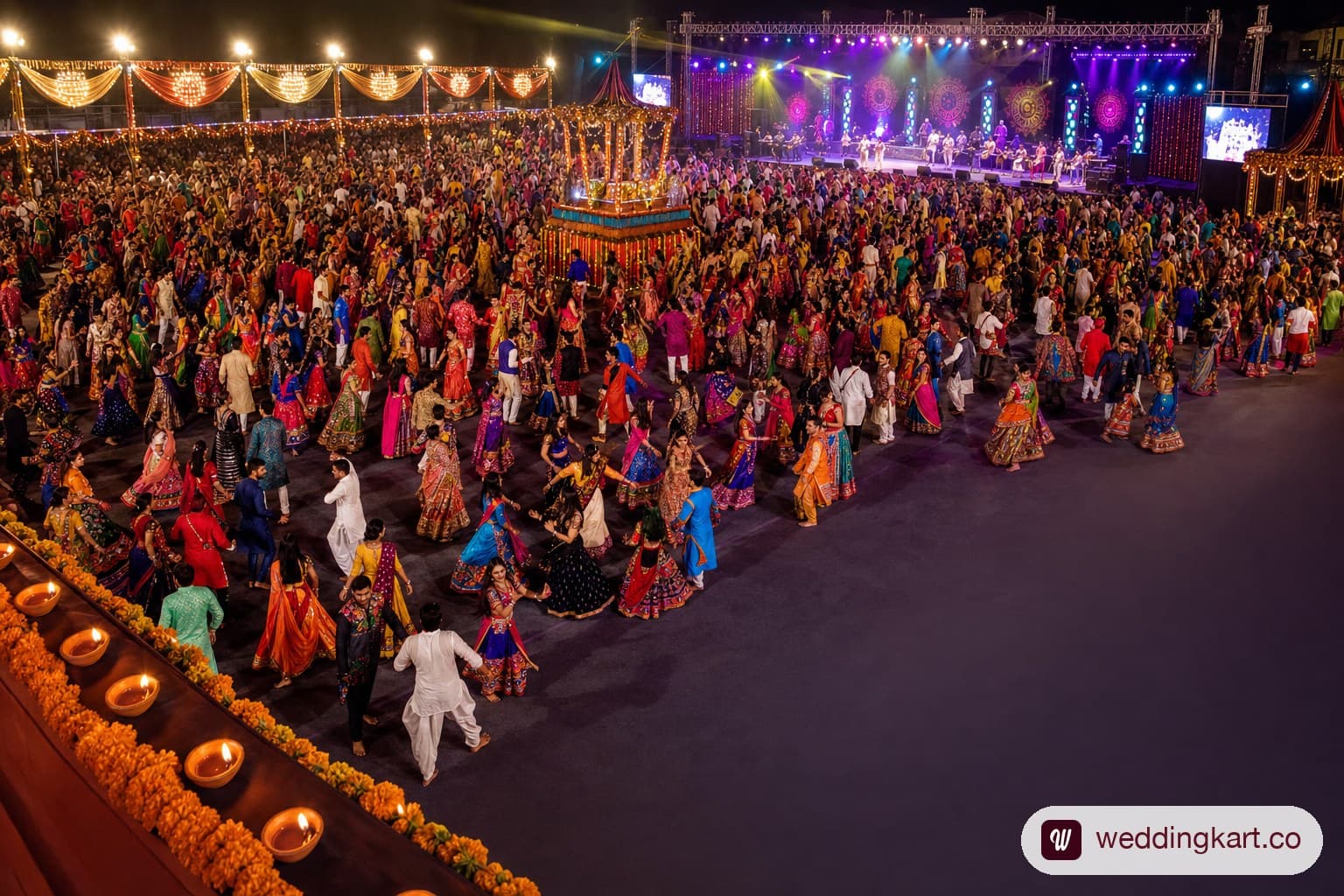A wide shot of a packed Garba night celebration at a large decorated hall in Ahmedabad, with hundreds of guests in vibrant chaniya choli outfits dancing in concentric circles, colourful stage lights, and a live orchestra