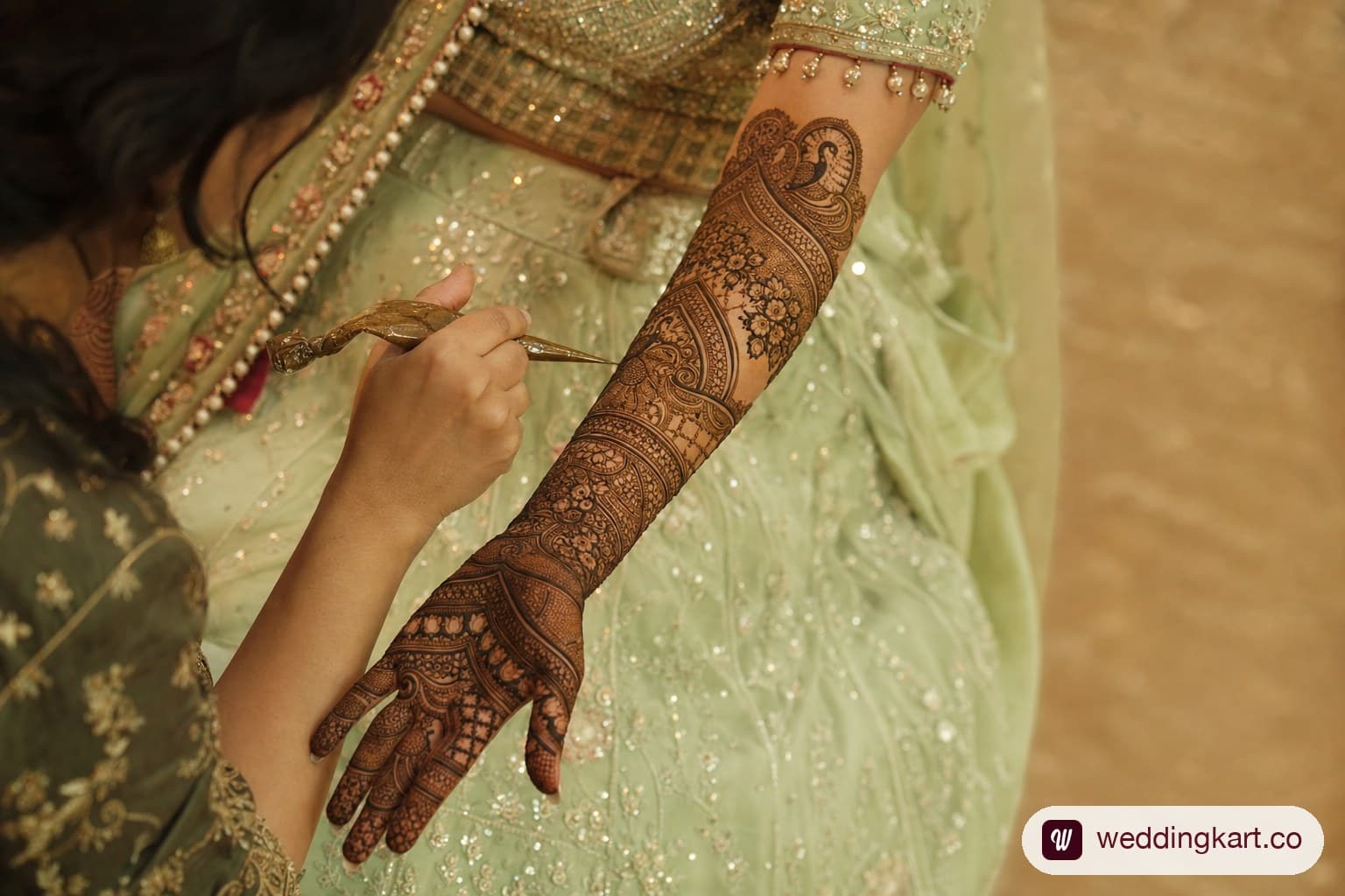 Close-up of a professional Mehendi artist applying intricate bridal Mehendi on a bride's hand and forearm with elaborate floral and peacock motifs, the bride wearing a light green chaniya choli