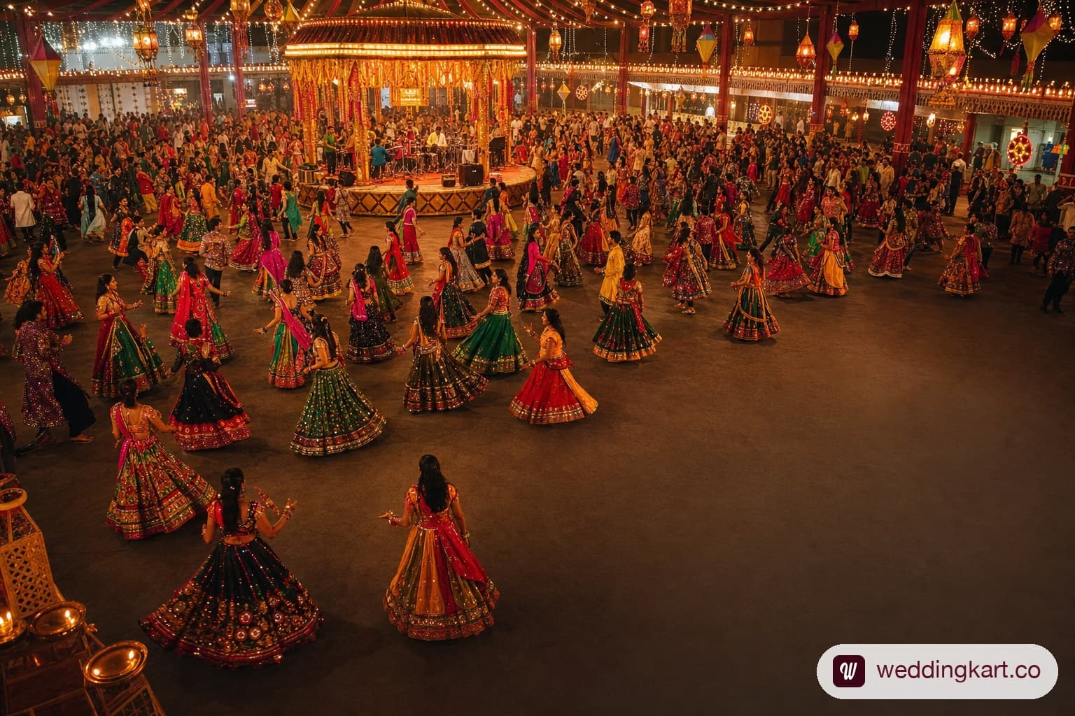 A high-energy Garba night at a large decorated hall in Ahmedabad, with hundreds of guests in vibrant chaniya choli outfits in red, green, orange, and fuchsia dancing in concentric circles, diyas and string lights illuminating the space, and a live Garba orchestra on the central stage